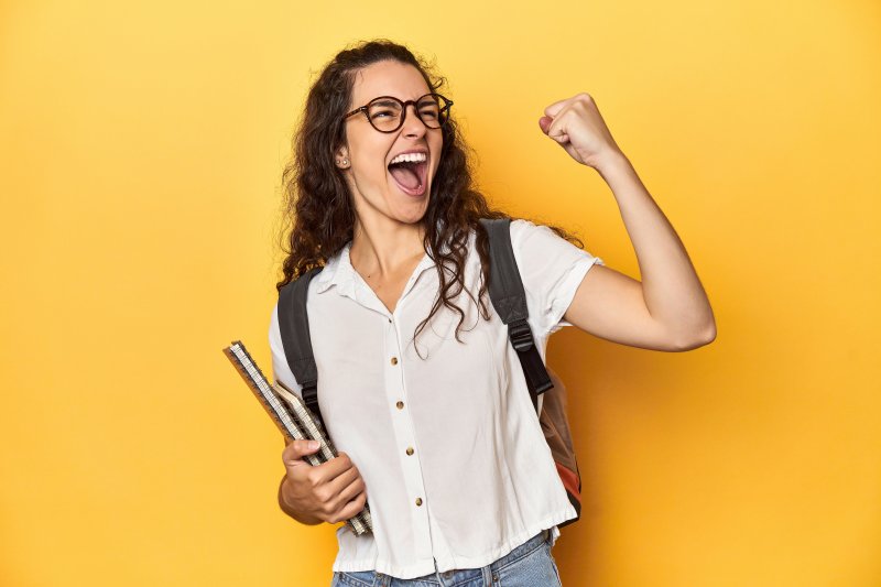 Young woman with glasses and wide smile fist pumping the air with one hand and carrying books in the other